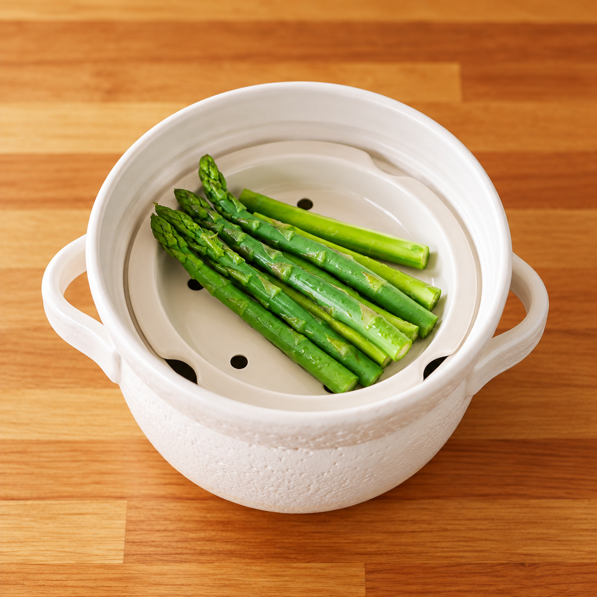Green asparagus arranged on a perforated ceramic steaming insert inside the white INAHO donabe.